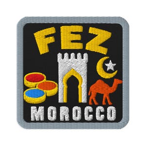 May include: A square embroidered patch with a grey border on a black background. It features the yellow text FEZ above a white tower, a yellow crescent moon and star, a brown camel, three colourful circular icons, and the white text MOROCCO.