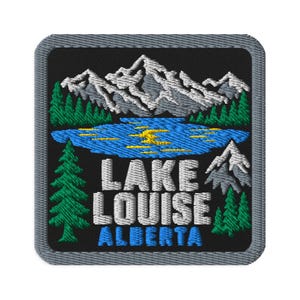 May include: A square embroidered patch featuring a black background with a landscape of grey mountains, a blue lake with yellow highlights, and green pine trees. The text LAKE LOUISE ALBERTA is embroidered in white and blue block letters.