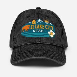 May include: A black, distressed baseball cap featuring a Salt Lake City, Utah graphic. The design includes a bison, mountains, a flower, and the sun. The text "SALT LAKE CITY UTAH" is in orange and white.