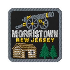 May include: A square embroidered patch with a black background and a gray border. It features a gray cannon with yellow accents, the white text MORRISTOWN, yellow text NEW JERSEY, a brown log cabin, and two green pine trees at the bottom.
