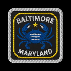 May include: Embroidered patch featuring a blue crab design, a star, and the words "BALTIMORE MARYLAND" in white. The patch has a black background with a yellow border and a silver edge.