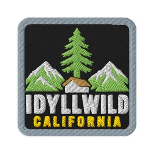 May include: A square embroidered patch with a black background and a light gray border. It features a green pine tree, two light green mountains with white peaks, a small brown cabin, and the white text IDYLLWILD above yellow text CALIFORNIA.