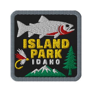 May include: A square embroidered patch with a gray border on a black background. It features a white fish with red gills, a yellow fishing fly, a green pine tree, and green mountains. The text reads ISLAND PARK in yellow and IDAHO in white.