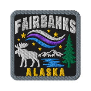 Op de afbeelding: Een vierkante geborduurde patch met een zwarte achtergrond en een grijze rand. Het bevat de witte tekst FAIRBANKS bovenaan en ALASKA onderaan. Het ontwerp toont een witte eland, een groene dennenboom, bergen, een blauw meer en gele sterren.