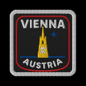 Op de afbeelding: Geborduurde patch met de tekst "WENEN" boven "OOSTENRIJK". Het ontwerp omvat een gele toren, een blauwe ster en een rode rand op een zwarte achtergrond. De patch heeft een witte rand.