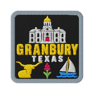 May include: A square embroidered patch with a grey border on a black background. It features a white building with a gold dome, the yellow text GRANBURY, white text TEXAS, a yellow longhorn, a pink flower, and a white sailboat on blue waves.