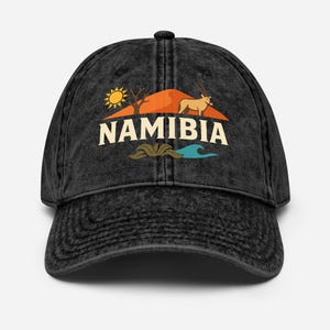 Op de afbeelding: Zwarte baseballcap met een vintage design. De cap heeft een landschapsafbeelding met een zon, bomen en een dier, met het woord "NAMIBIA" in witte letters. De cap heeft een gebogen klep en een verstelbare band.