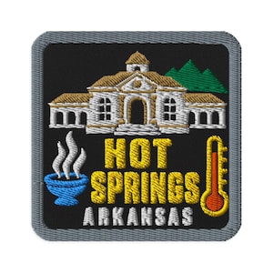 Op de afbeelding: Een vierkante geborduurde patch met een grijze rand op een zwarte achtergrond. Het toont een wit gebouw met een beige dak, groene bergen, een blauwe dampende kom, een gele thermometer en de tekst HOT SPRINGS ARKANSAS in geel en wit.