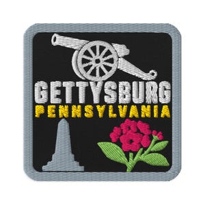 Puede incluir: Un parche bordado cuadrado con borde gris sobre fondo negro. Presenta un cañón blanco, el texto blanco GETTYSBURG, el texto amarillo PENNSYLVANIA, la silueta de un monumento gris y un grupo de flores rosas con hojas verdes.