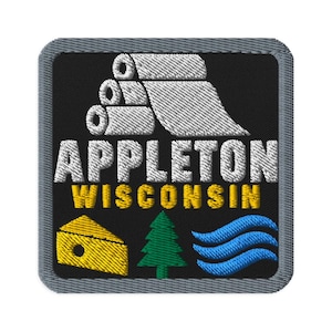 May include: A square embroidered patch with a black background and a gray border. It features white paper rolls, the text APPLETON WISCONSIN in white and yellow, a yellow wedge of cheese, a green pine tree, and blue wavy lines.