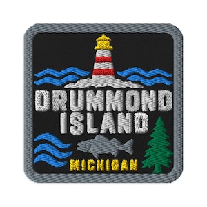 May include: A square embroidered patch with a black background and a grey border. It features a lighthouse with red and white stripes, blue waves, a grey fish, a green pine tree, and the white text DRUMMOND ISLAND with yellow text MICHIGAN.