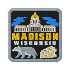 Puede incluir: Un parche bordado cuadrado con fondo negro que muestra un edificio del capitolio en blanco y dorado, el texto MADISON WISCONSIN en amarillo y blanco, un tejón gris y un trozo de queso blanco con puntos amarillos, bordeado en gris claro.