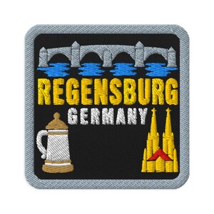 Puede incluir: Un parche bordado cuadrado con fondo negro que muestra un puente de piedra gris sobre agua azul, el texto amarillo REGENSBURG GERMANY, una jarra de cerveza blanca con tapa marrón y una catedral amarilla con techo rojo en la parte inferior derecha.