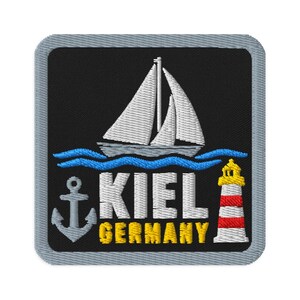 Puede incluir: Un parche bordado cuadrado con fondo negro y borde gris. Presenta un velero blanco sobre olas azules, un ancla gris, un faro rojo y blanco, y el texto blanco KIEL con el texto amarillo GERMANY debajo.