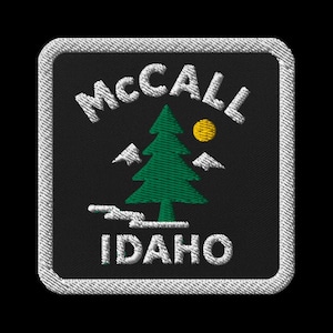 May include: Embroidered patch featuring the text "McCall Idaho" in white, with a green pine tree, a yellow sun, and white mountain silhouettes on a black background. The patch has a white border.