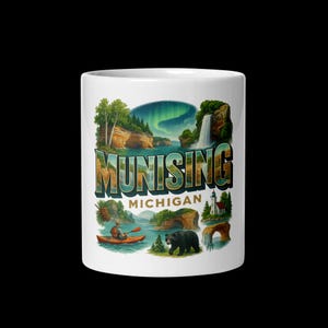 May include: White ceramic mug featuring a colorful illustration of Munising, Michigan. The design includes the city's name, scenic landscapes, a waterfall, a bear, and a kayaker. The mug is a standard size for hot beverages.