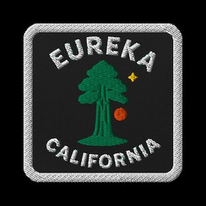 Op de afbeelding: Geborduurde patch met de witte woorden "EUREKA CALIFORNIA", gebogen rond een groene boom, een gele ster en een oranje cirkel op een zwarte achtergrond. De patch heeft een witte rand.