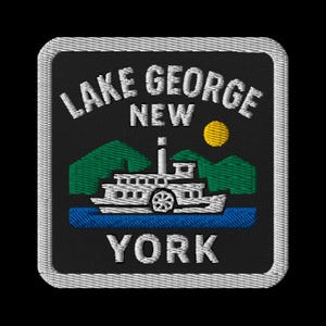 May include: Embroidered patch featuring a steamboat on blue water, with green mountains and a yellow sun. The text "LAKE GEORGE NEW YORK" is arched above the image in white, with a gray border.