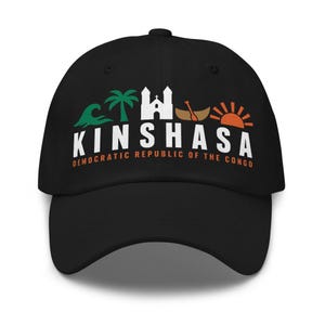 Può includere: Un cappellino da baseball nero con un design grafico che include un'onda verde, una palma, un edificio bianco, una barca marrone con un remo e un sole arancione. Il testo riporta KINSHASA e DEMOCRATIC REPUBLIC OF THE CONGO in bianco e arancione.