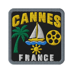 May include: A square embroidered patch with a black background and a grey border. It features the yellow text CANNES above a green palm tree, a white sailboat on blue waves, a yellow film reel, a yellow sun, and the white text FRANCE.