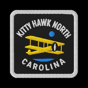 May include: Embroidered patch featuring a yellow biplane, a blue dot, and a blue line, set against a black background. The text "KITTY HAWK NORTH CAROLINA" is arched around the biplane in white. The patch has a white border.