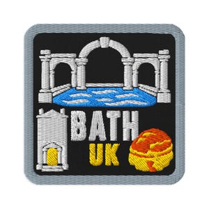 Puede incluir: Un parche bordado cuadrado con borde gris sobre fondo negro. Presenta un arco de estilo romano blanco sobre una piscina azul, un edificio blanco con puerta amarilla, el texto blanco BATH, el texto amarillo UK y un bollo redondo naranja y amarillo.