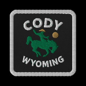 May include: Embroidered patch featuring the words "Cody Wyoming" in white, arched over a green silhouette of a cowboy riding a horse. A gold-colored ball is to the right of the cowboy. The patch has a black background and a white border.