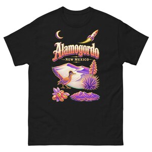 Peut inclure: Un t-shirt noir avec un graphisme coloré représentant un grand géocoucou, des dunes de désert, une fusée, un croissant de lune et des montagnes. Le texte indique Alamogordo New Mexico dans une police stylisée aux tons violets, orange et jaunes.
