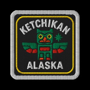 May include: Embroidered patch featuring a stylized totem owl design in green, red, and black. The text "KETCHIKAN ALASKA" is embroidered in white, with a yellow border and a silver-gray edge. The patch is square-shaped.