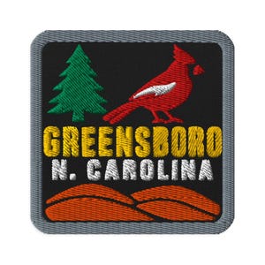 May include: A square embroidered patch with a black background and a grey border. It features a green pine tree, a red cardinal bird, and orange hills. The text reads GREENSBORO in yellow and N. CAROLINA in white, all stitched in thread.