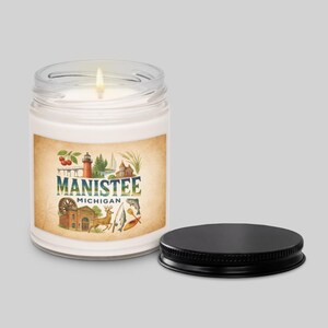 Op de afbeelding: Een brandende witte kaars in een heldere glazen pot met een zwart deksel. Het etiket toont de tekst "Manistee Michigan" met illustraties van een vuurtoren, gebouwen en dieren in het wild. De pot staat op een lichtgrijze achtergrond.