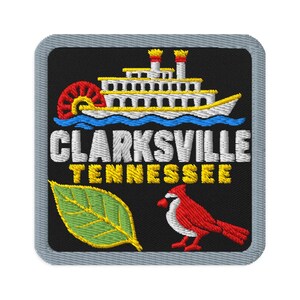 Op de afbeelding: Een vierkante geborduurde patch met een grijze rand op een zwarte achtergrond. Het toont een witte raderstoomboot, de tekst CLARKSVILLE TENNESSEE in wit en geel, een groen blad en een rode kardinaalvogel met een witte borst.