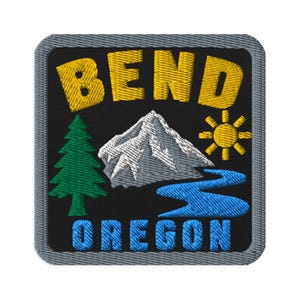 Może przedstawiać: Kwadratowa haftowana naszywka z czarnym tłem i szarą obwódką. Napis BEND jest żółty u góry, a OREGON niebieski u dołu. Wzór przedstawia zieloną sosnę, białą ośnieżoną górę oraz niebieską rzekę umieszczoną w centralnej części naszywki.