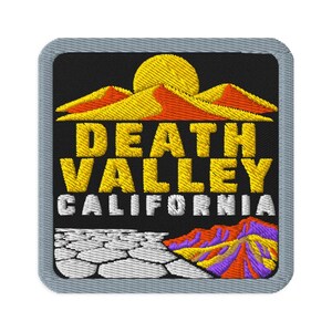 Könnte beinhalten: Ein quadratischer gestickter Aufnäher mit grauem Rand auf schwarzem Hintergrund. Er zeigt eine gelbe Sonne über orangefarbenen und gelben Sanddünen, den gelben Schriftzug DEATH VALLEY, weißen Text CALIFORNIA, einen weißen rissigen Wüstenboden und violette Berge.