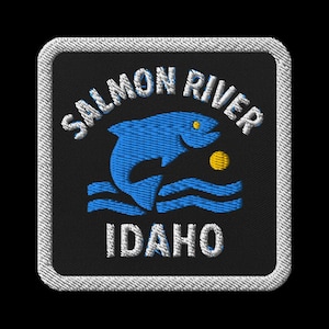 Puede incluir: Parche bordado con un pez azul saltando sobre líneas onduladas y un sol amarillo. Las palabras "SALMON RIVER" e "IDAHO" están en blanco, arqueadas arriba y abajo del pez. El parche tiene un fondo negro y un borde blanco cosido.