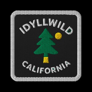 May include: Embroidered patch with the words "IDYLLWILD CALIFORNIA" in white, arched around a green pine tree and yellow sun on a black background. The patch has a white border.