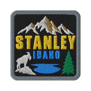 May include: A square embroidered patch with a black background and a gray border. It features white and tan mountain peaks, the yellow text STANLEY, blue text IDAHO, a white mountain goat, a blue lake, and a green pine tree.