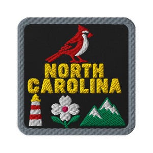 Puede incluir: Un parche bordado cuadrado con borde gris sobre fondo negro. Presenta un pájaro cardenal rojo sobre el texto amarillo NORTH CAROLINA. Debajo del texto hay un faro rojo y blanco, una flor de cornejo blanca y unas montañas de color verde.
