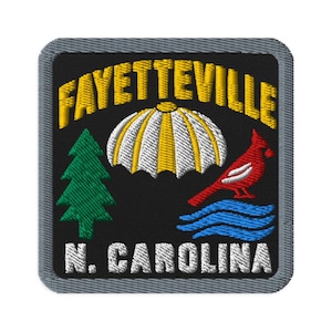 Puede incluir: Un parche bordado cuadrado con fondo negro y borde gris. Presenta el texto amarillo FAYETTEVILLE en la parte superior y N. CAROLINA en la inferior. El centro muestra un paracaídas amarillo y blanco, un pino verde, un pájaro rojo y olas azules.