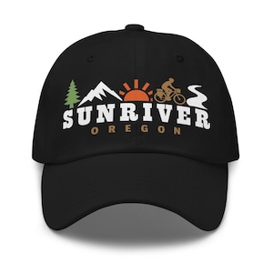 May include: A black baseball cap featuring a graphic with a green pine tree, white mountain peak, orange sun, a cyclist on a bicycle, and a white winding path. The text reads SUNRIVER in white and OREGON in brown below the graphic elements.