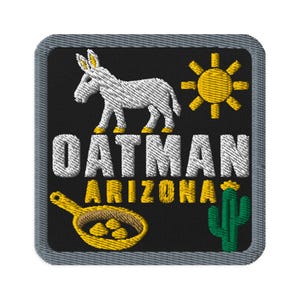 Puede incluir: Un parche bordado cuadrado con fondo negro y borde gris. Presenta un burro blanco, un sol amarillo, una sartén amarilla con tres elementos amarillos y un cactus verde. El texto OATMAN ARIZONA está bordado en color blanco y amarillo.
