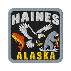 Op de afbeelding: Een vierkante geborduurde patch met een grijze rand op een zwarte achtergrond. Het bevat de witte tekst HAINES bovenaan en ALASKA onderaan in het geel. Het ontwerp bevat een witte adelaar, een grijze beer en een oranje vis.
