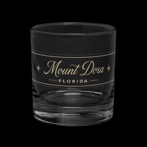 May include: A clear, lowball glass with a black band featuring the text "Mount Dora FLORIDA" in a gold script font. The glass has a thick base and is designed for serving beverages.