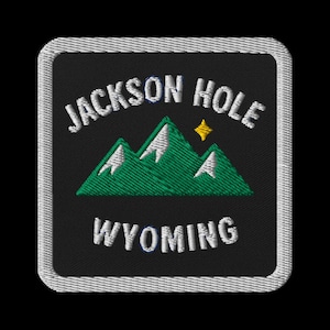 May include: Embroidered patch featuring the text "JACKSON HOLE WYOMING" in white, arched above a green mountain range with white peaks and a yellow star. The patch has a black background and a white stitched border.