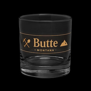 May include: A clear, lowball glass with a black exterior and a gold-coloured band featuring the word "Butte" and "Montana". The design includes a pickaxe and a mountain graphic.