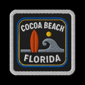 May include: Embroidered patch featuring "COCOA BEACH FLORIDA" in white text. The design includes an orange surfboard, a yellow sun, and a gray wave on a black background, framed by blue, yellow, and white borders.