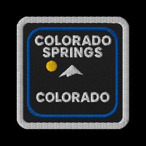 May include: Embroidered patch featuring the text "COLORADO SPRINGS COLORADO" in white, with a yellow sun and a white mountain graphic on a black background. The patch has a blue border and a white stitched edge.