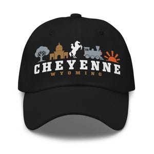 Könnte beinhalten: Eine schwarze Baseballkappe mit einem Grafikdruck, der einen grauen Baum, ein beiges Gebäude, ein weißes Pferd mit Reiter, einen grauen Zug und eine orangefarbene Sonne zeigt. Darunter steht der Text CHEYENNE in Weiß und WYOMING in Beige.