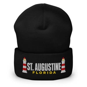 Könnte beinhalten: Eine schwarze Mütze mit Umschlag, die eine Stickerei von zwei rot-weiß gestreiften Leuchttürmen zeigt, welche den weißen Schriftzug ST. AUGUSTINE und den gelben Schriftzug FLORIDA auf der Vorderseite des Umschlags einrahmen.
