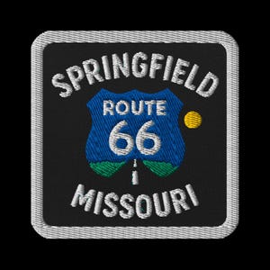 Puede incluir: Parche bordado con las palabras "Springfield Missouri" y "Route 66". El diseño incluye un escudo azul con el número "66" y un punto amarillo. El parche tiene un fondo negro y un borde blanco.